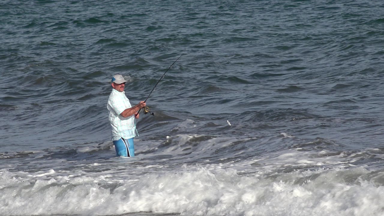 The fisherman remains waist-deep in Los Cabos water while a small wave lifts behind him and white foam spreads across the lower frame.