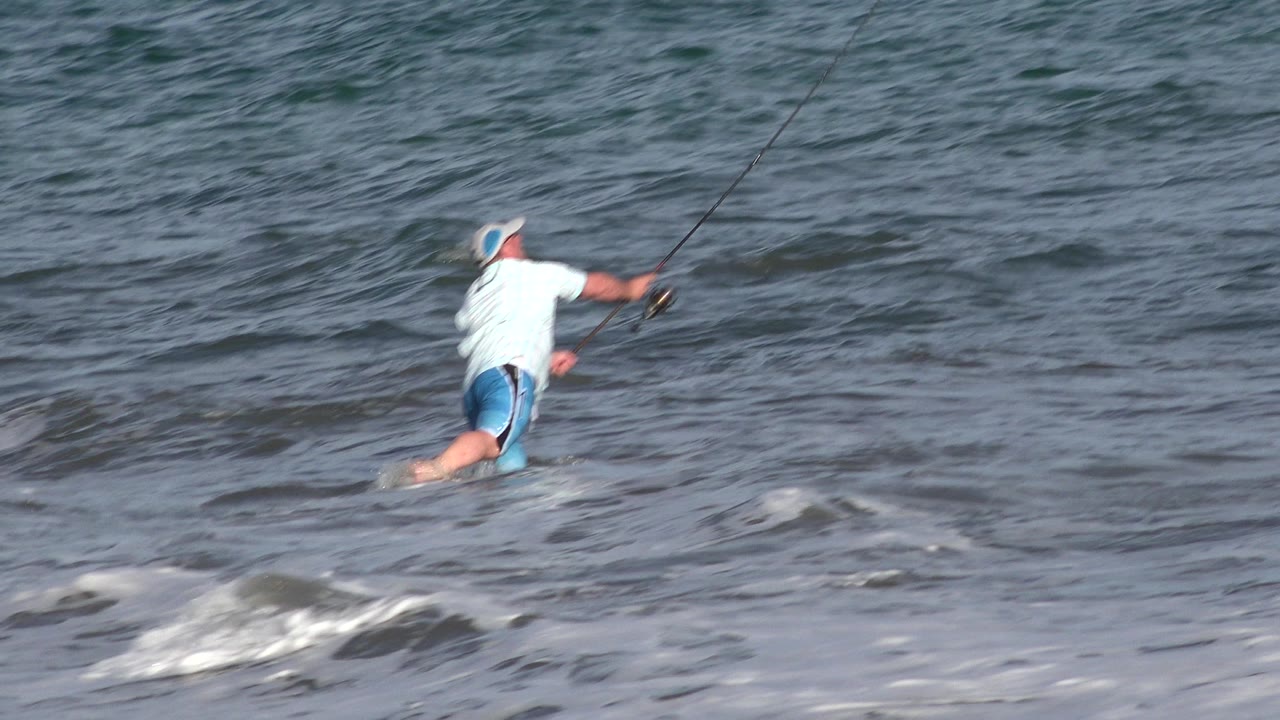 A fisherman in Los Cabos, Mexico wades knee-deep into choppy shallow water with his rod angled forward as small waves break around his legs.