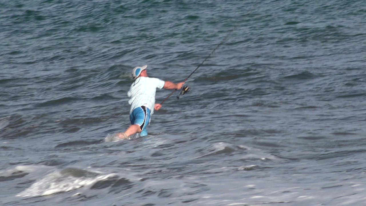 The Los Cabos fisherman steps through foamy surf with one leg lifted and the fishing rod extending out over the water.