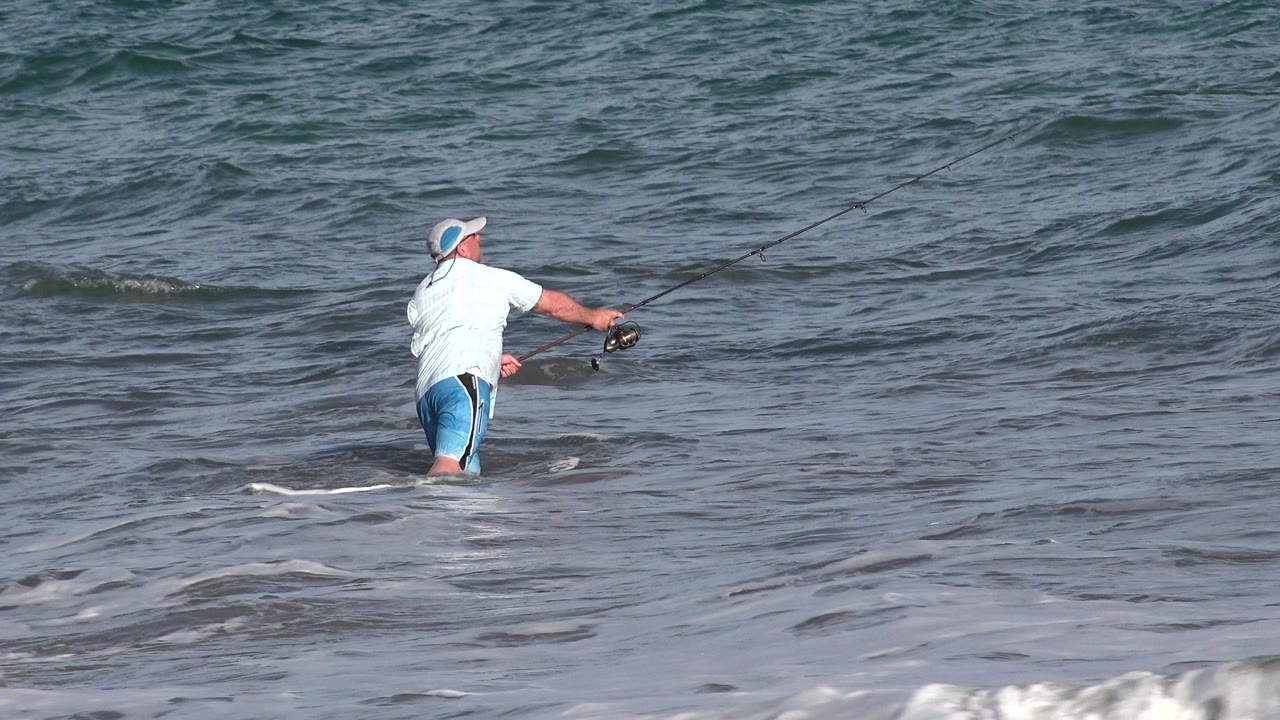Fisherman standing in surf with rod extended toward open water.