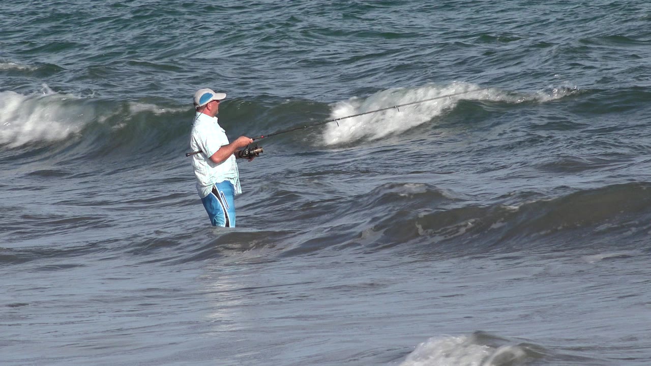 The angler remains in the Los Cabos shoreline water with the rod angled forward while a white-capped wave breaks across the background.