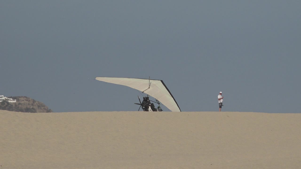 At 0:01 in Los Cabos, Mexico, the hang glider remains centered behind the dune line while the man stands at the right against empty sky.