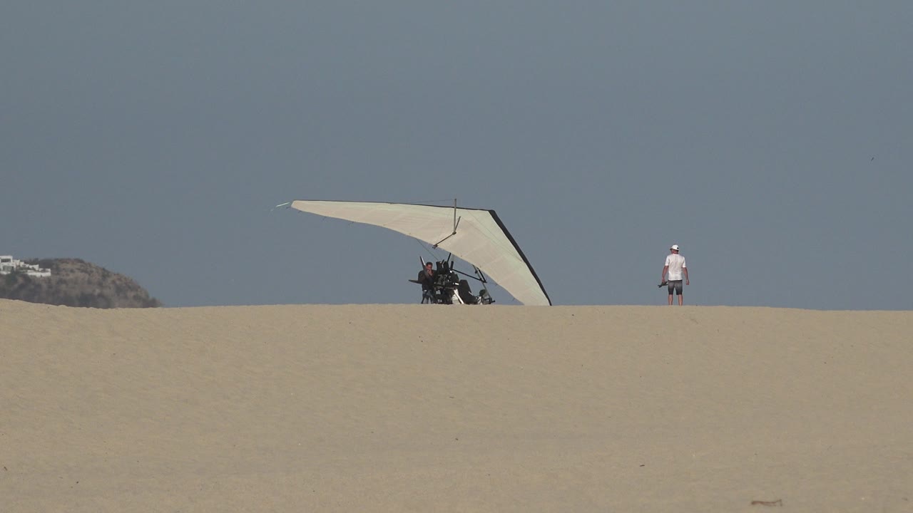 A hand glider rests on a Los Cabos sand ridge with distant people beside it and one person standing farther right under a clear blue sky.