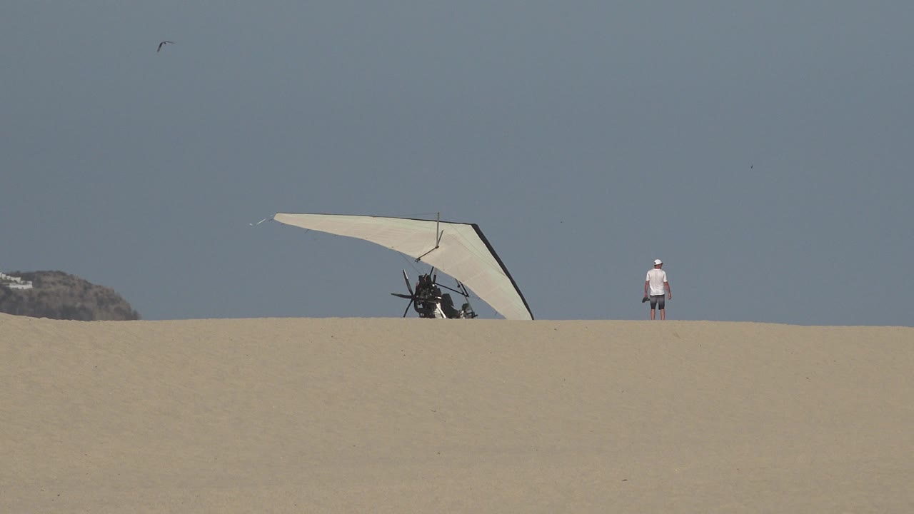White hang glider on a sandy ridge with a distant person at right.