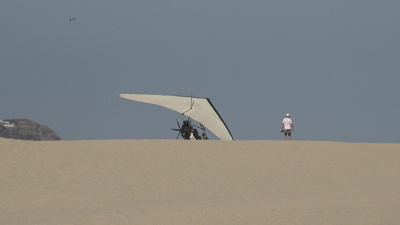 At 0:01, the hang glider remains partly behind the sandy crest in Los Cabos, Mexico, while the distant person stands separated on the right side of the frame.