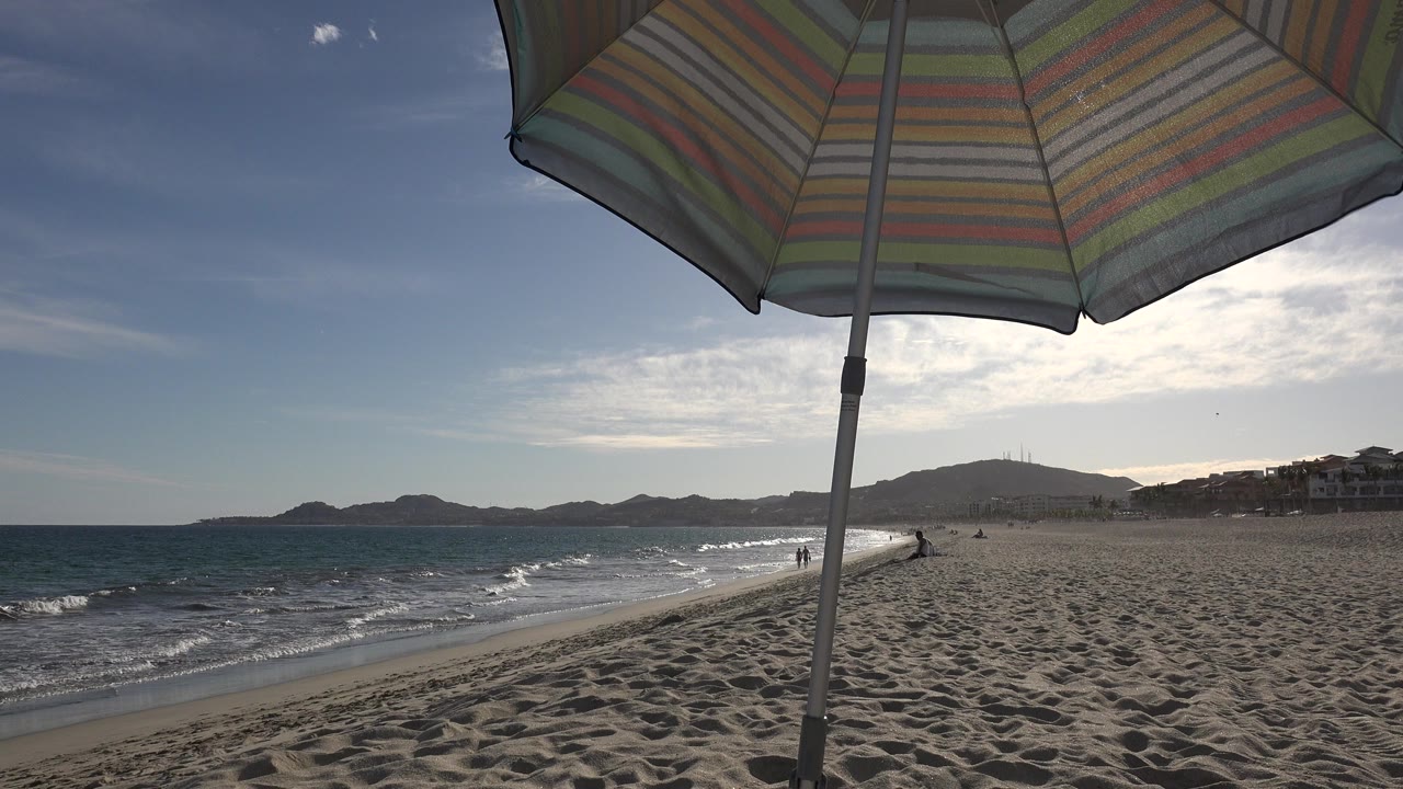 Striped beach umbrella over a sandy shoreline with waves and distant hills.