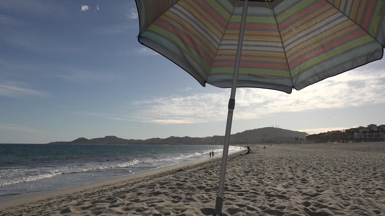 Striped beach umbrella and pole dominate the foreground on a Los Cabos beach, with low waves, distant hills, shoreline buildings, and small beachgoers in the background.