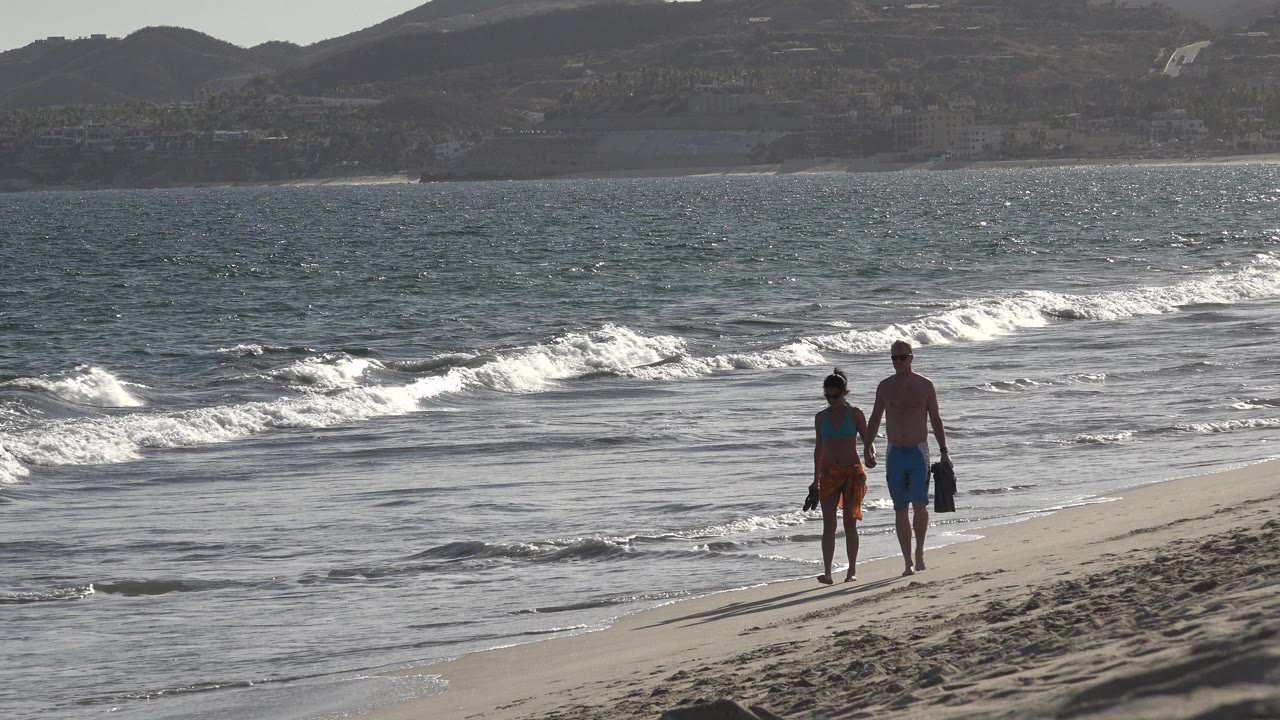 At the start of the Los Cabos clip, a man and woman walk hand in hand on wet sand while white surf breaks behind them and hills sit across the bay.