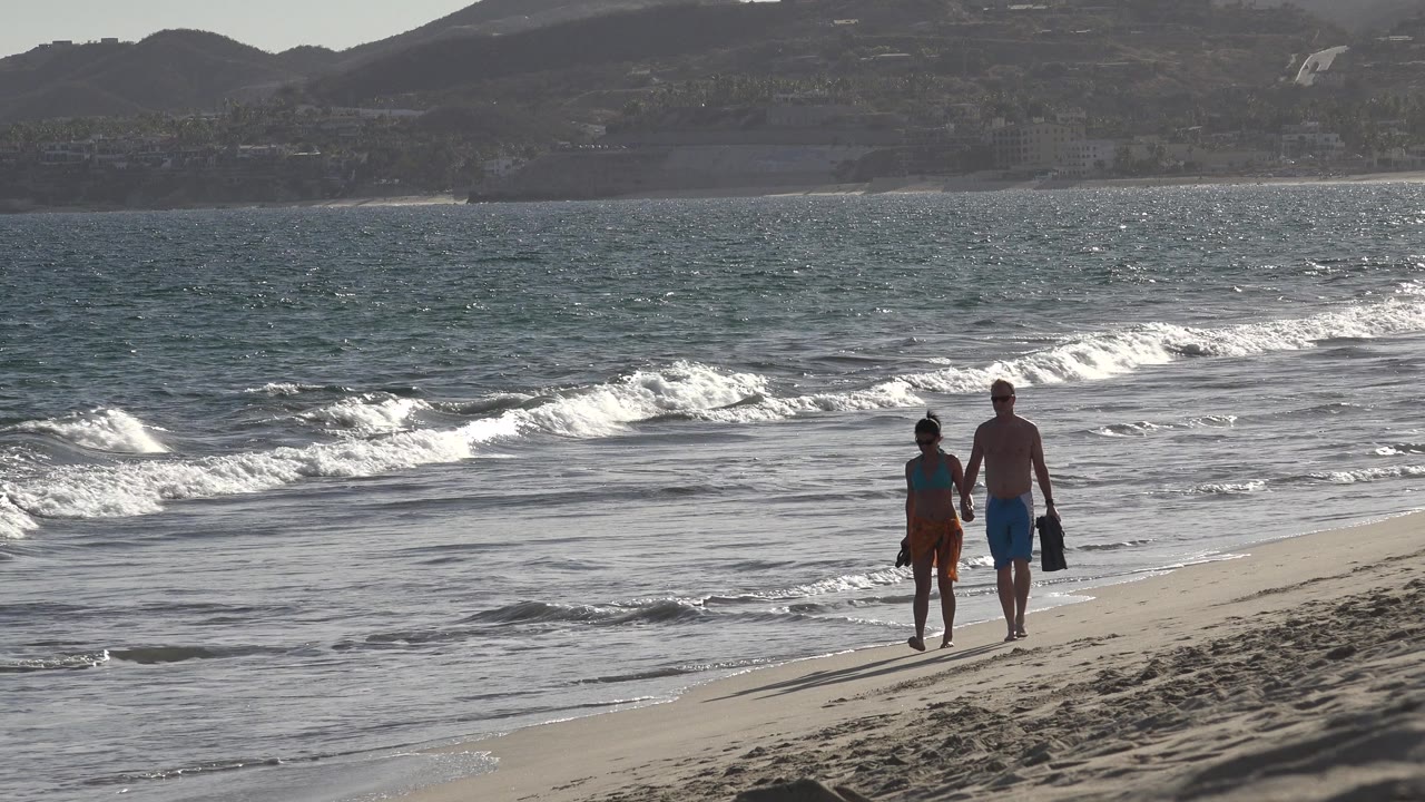 One second in, the couple remains near the waterline in Los Cabos as a bright band of foam crosses the midground and the beach curves to the right.