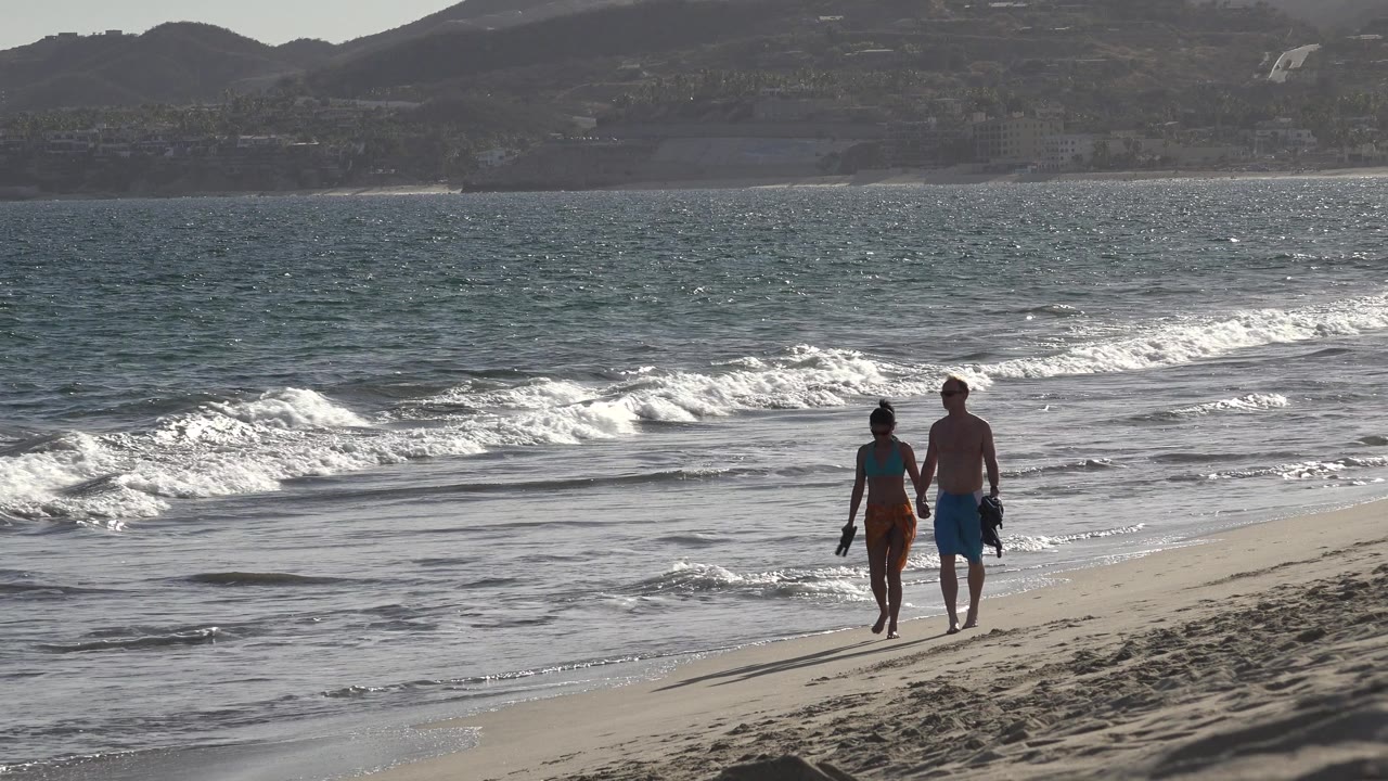 Couple walking together on a sunlit beach with ocean surf behind.
