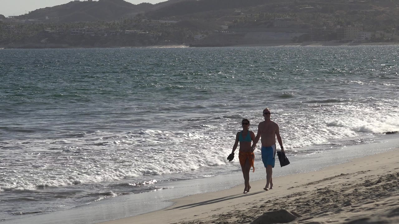 Couple walking hand in hand along a sunny beach with waves behind them.