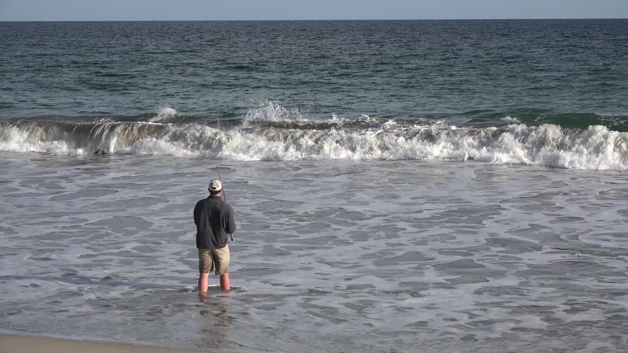 Man fishing in shallow surf facing the ocean.