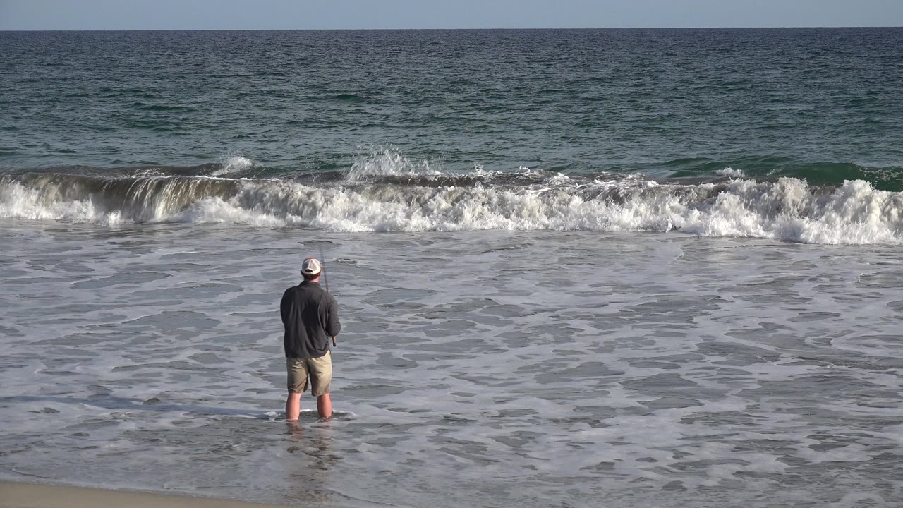 The fisherman remains centered near the waterline in Los Cabos, Mexico, while a white band of surf breaks across the midground.
