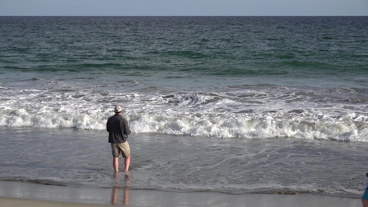 The fisherman remains centered at the Los Cabos waterline with white foam spreading across the wet sand and darker blue water beyond.