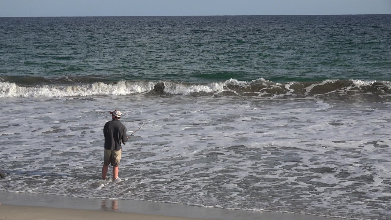 The fisherman stands closer to center with his back to camera in Los Cabos, Mexico, while a low line of waves rolls toward the wet beach.