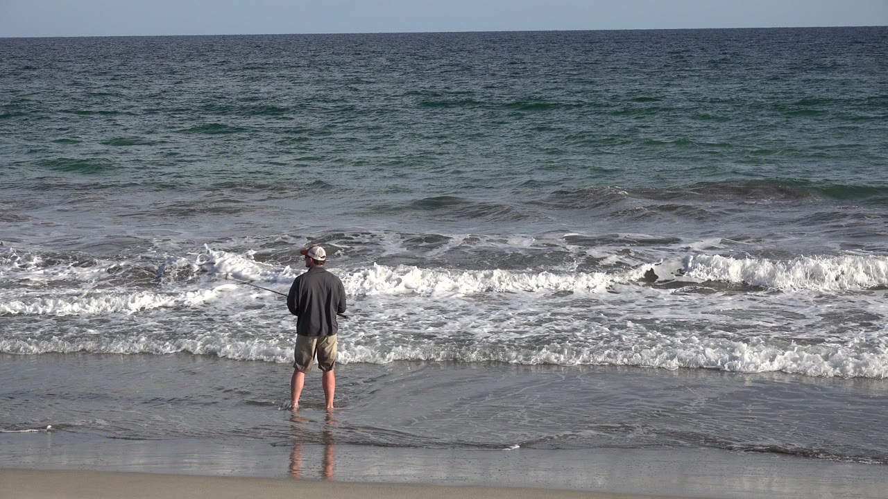In Los Cabos, Mexico, a man in a baseball hat and cargo shorts stands barefoot on wet sand facing the ocean as a low wave breaks in front of him.