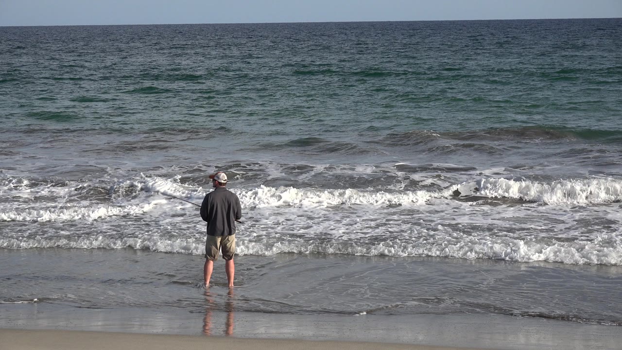 The fisherman remains centered on the Los Cabos shoreline, holding a fly rod while white foam spreads across the shallow water at his feet.