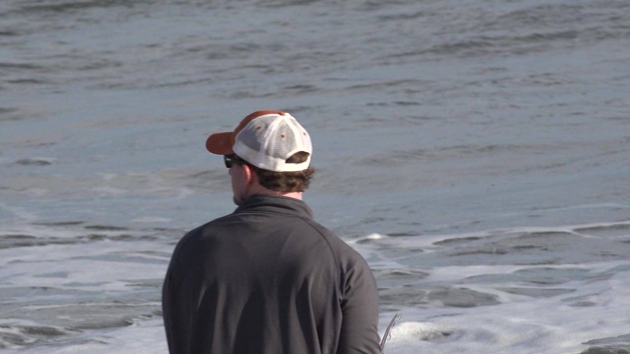 In Los Cabos, Mexico, a man seen from behind stands near the waterline with a fishing rod as gray-blue surf rolls across the frame.