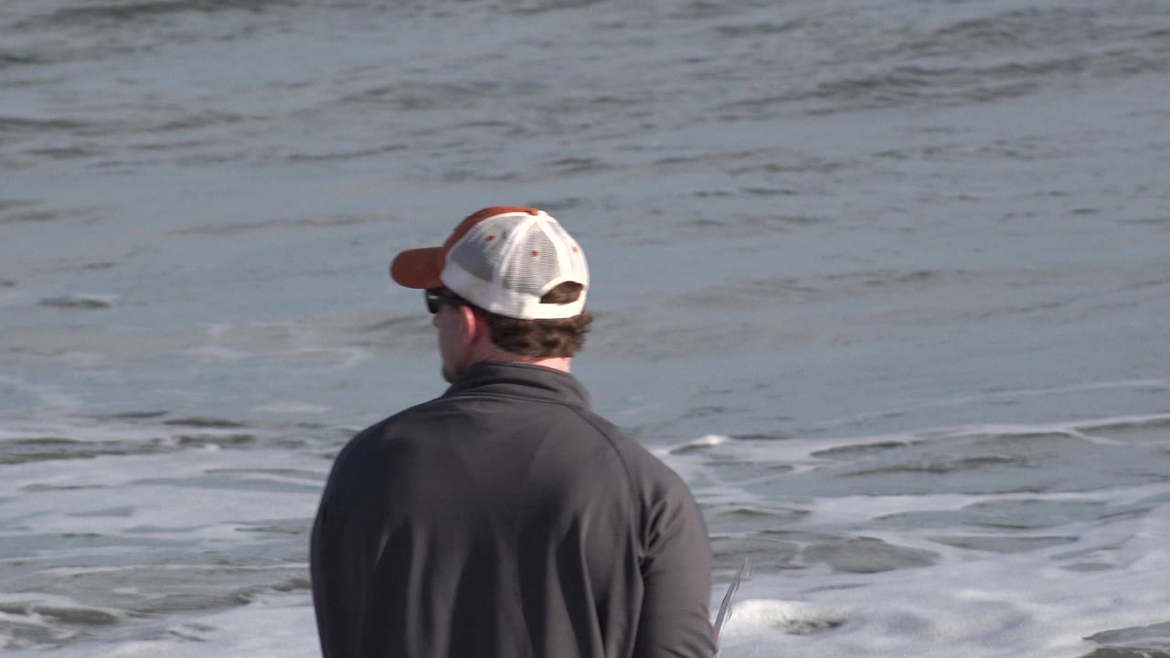 The man remains centered from behind in Los Cabos, Mexico, with white foam spreading across the water in front of his fishing position.