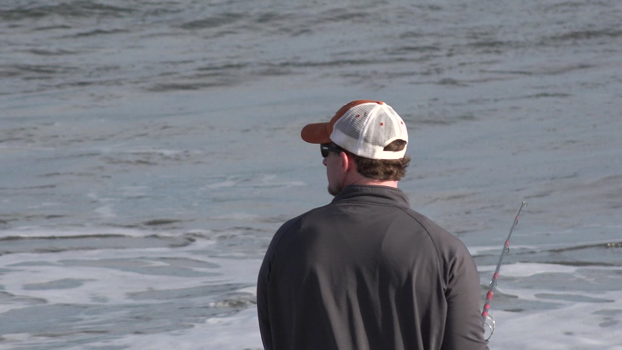 Angler in cap watches the surf with rod at right.