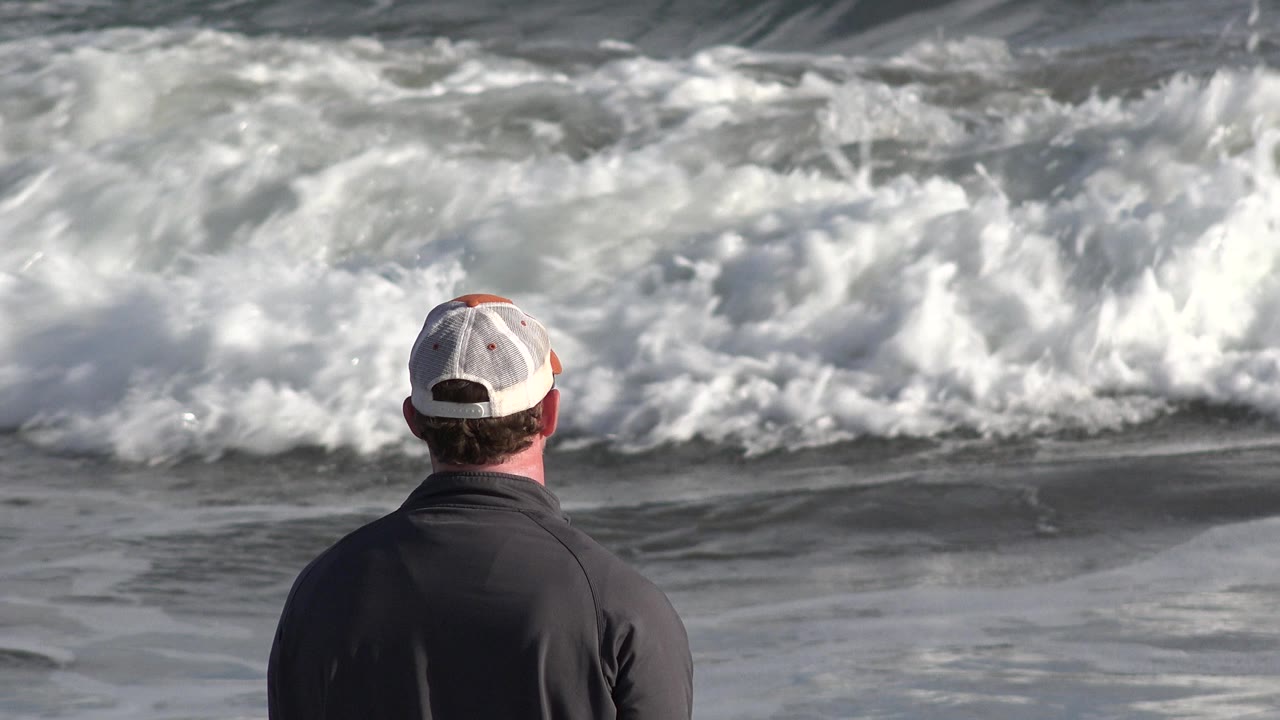 At 0:00 in Los Cabos, Mexico, a man in a light baseball cap and dark shirt stands with his back to camera as a white wave breaks across the upper frame.