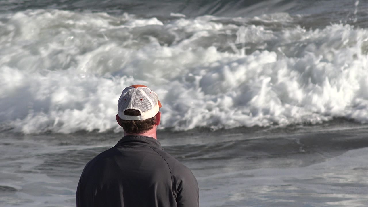 At 0:01 in Los Cabos, Mexico, the man's cap and shoulders remain centered while foamy surf stretches wider behind him across the gray water.