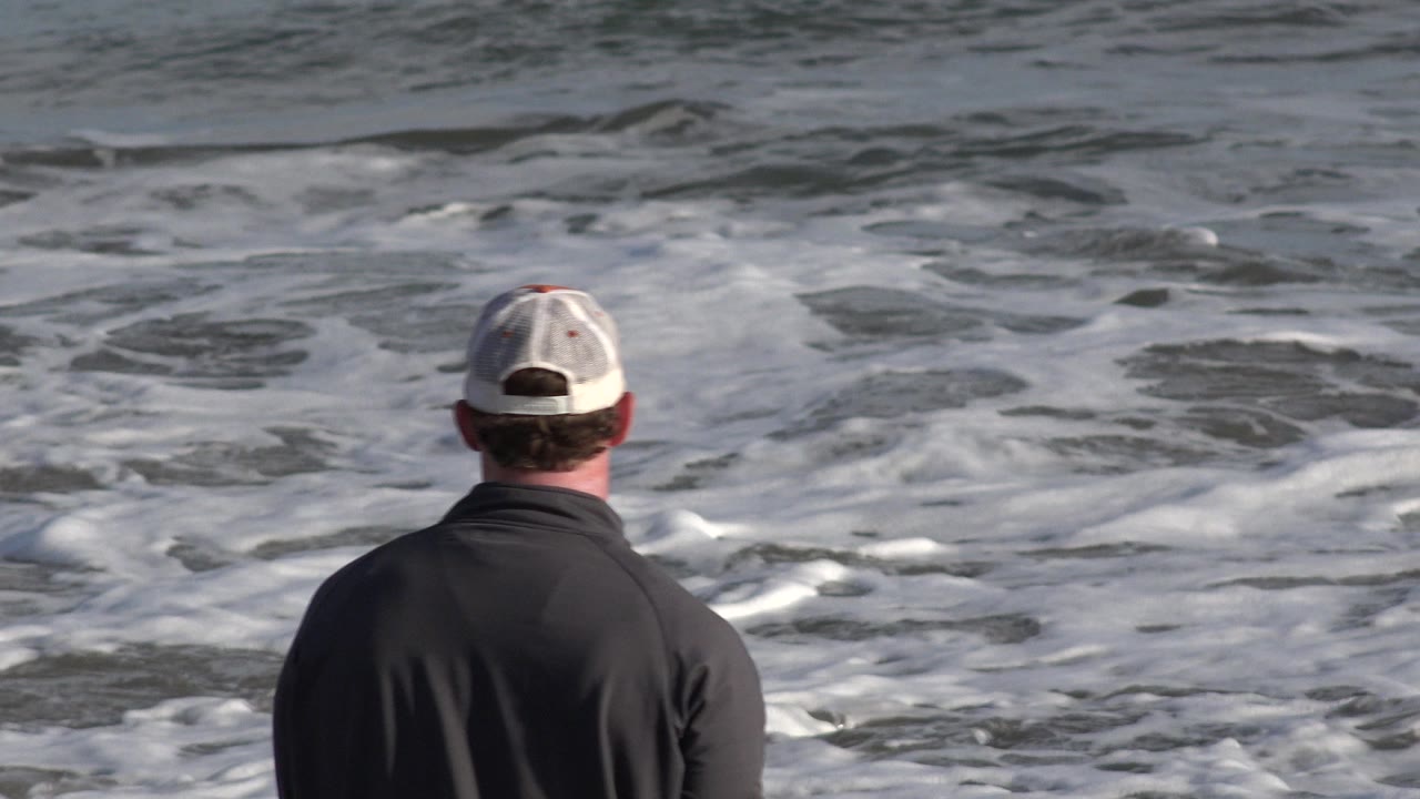 A man in a light cap and dark shirt stands with his back to camera facing foamy surf in Los Cabos, Mexico at the start of the clip.