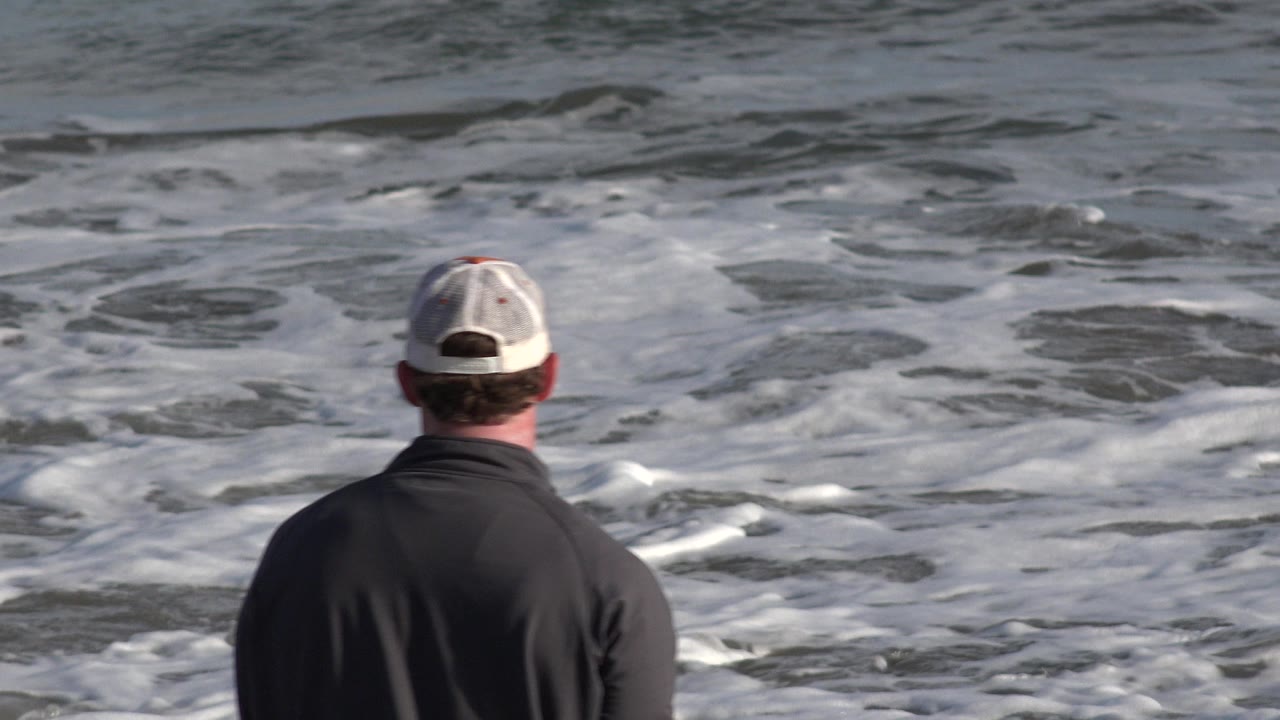 The man remains centered from behind as white foam spreads across the gray-blue water in Los Cabos, Mexico.