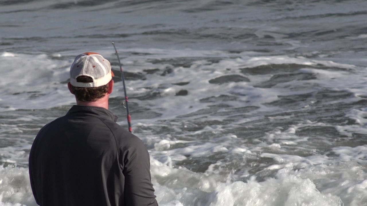 Man seen from behind fishing beside foamy surf.