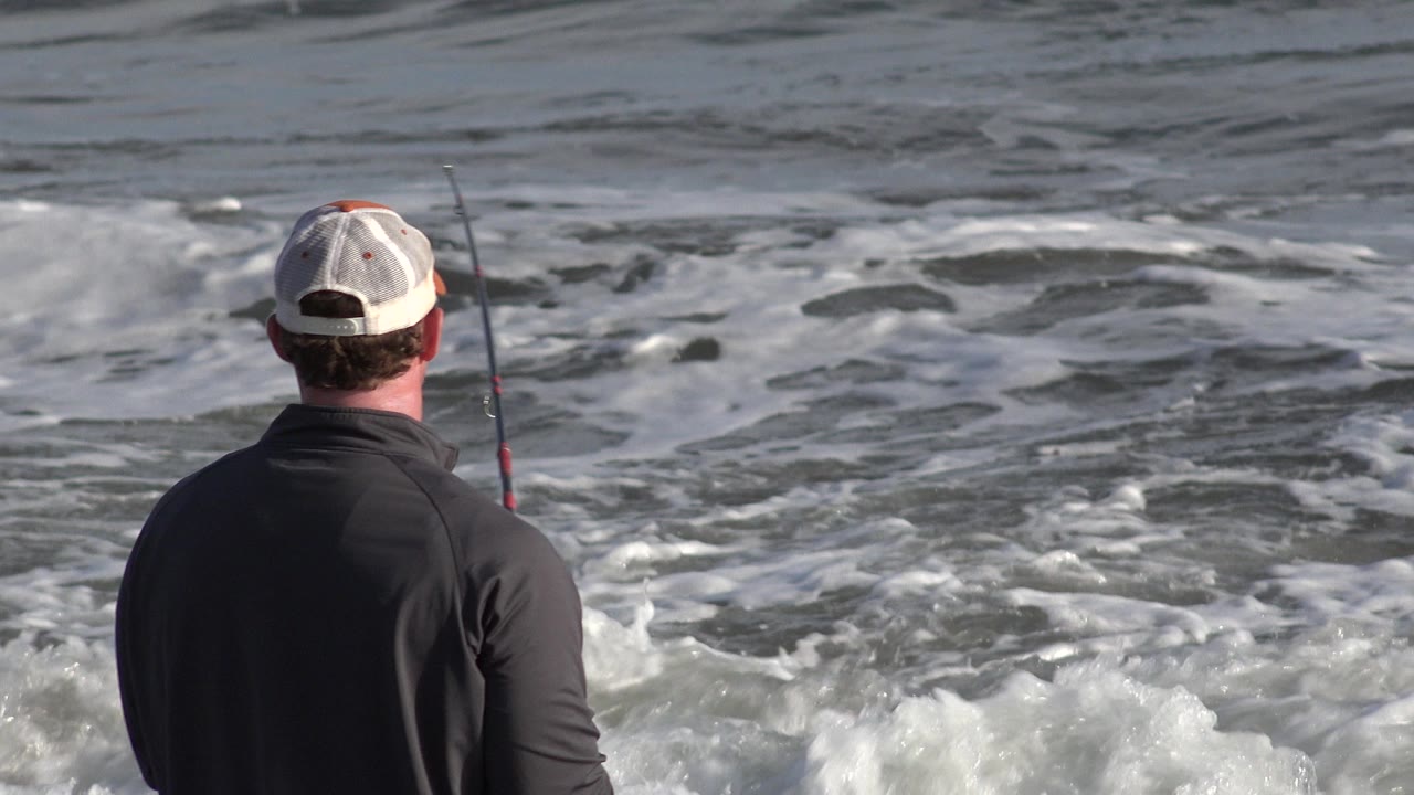 The fisher remains centered from behind in Los Cabos, Mexico, with the rod held near his right side and broken white foam spreading across the sea.