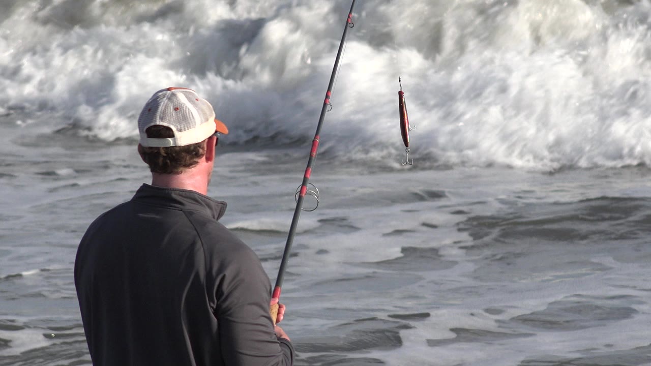 Man seen from behind holding a fishing rod near breaking surf.