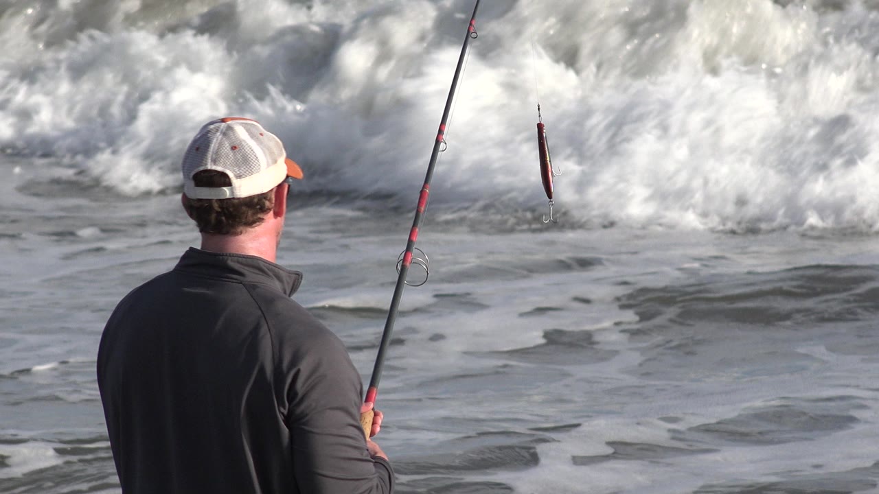 At 0:01 in Los Cabos, Mexico, the fisherman remains back-facing with his rod upright as white surf rolls across the beach in front of him.