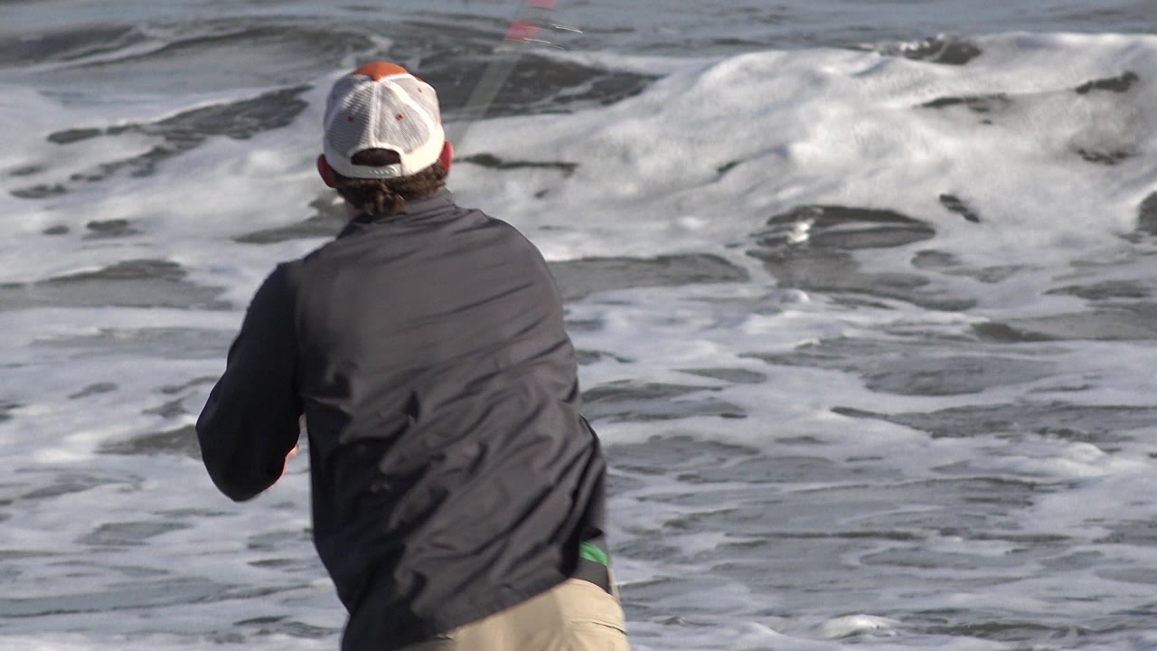 Man from behind fishing beside foamy surf.