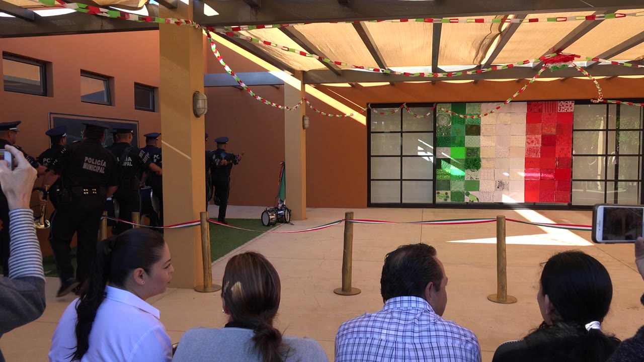 Uniformed police move across the left midground in San Jose del Cabo while seated viewers face a decorated wall with red, white, and green panels.