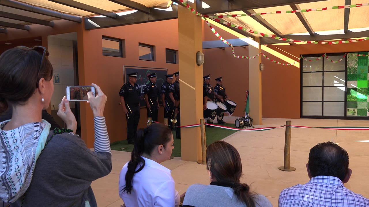 In San Jose del Cabo, spectators sit and stand in the foreground while uniformed police gather with drums near an orange-walled performance area.