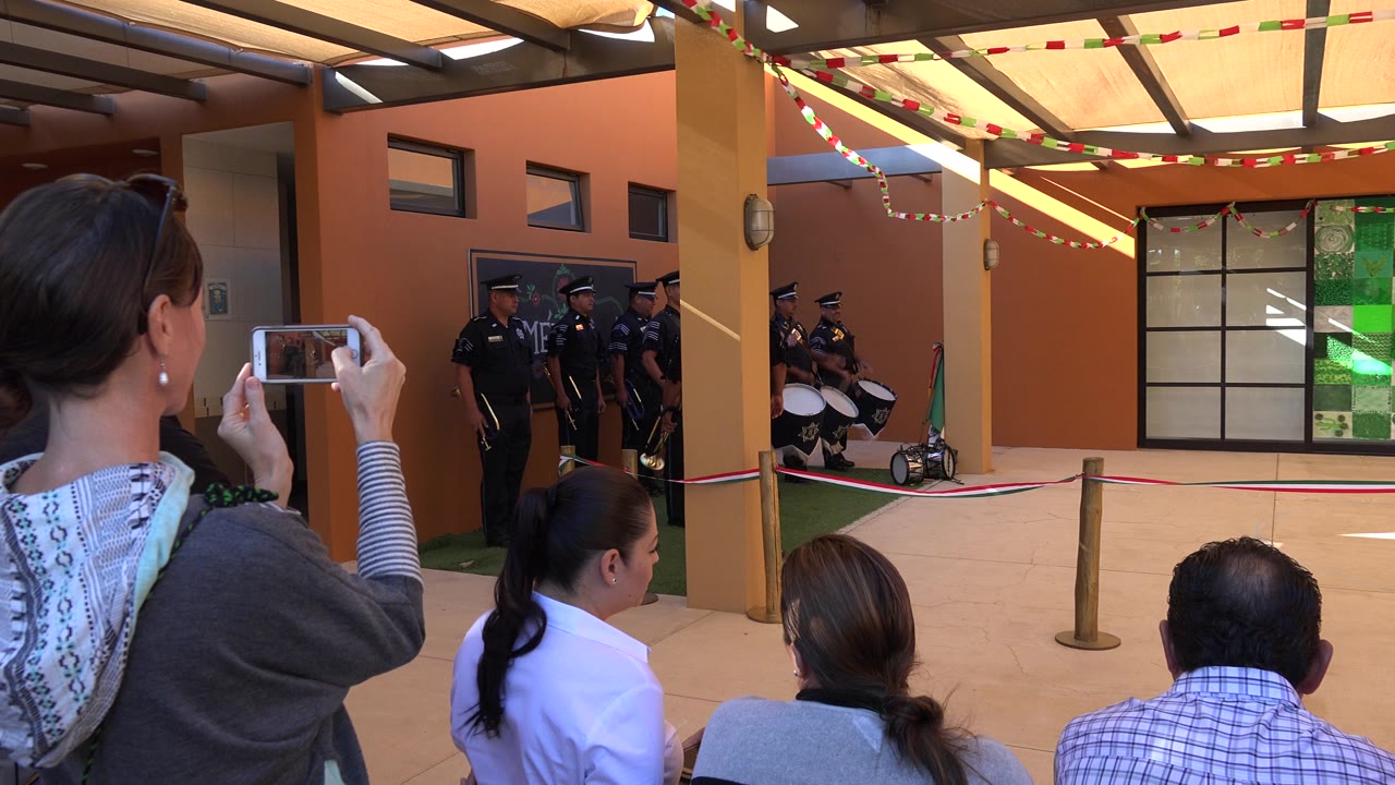 In San Jose del Cabo, seated spectators and a woman filming on a phone face several uniformed police officers positioned by drums under a shaded canopy.