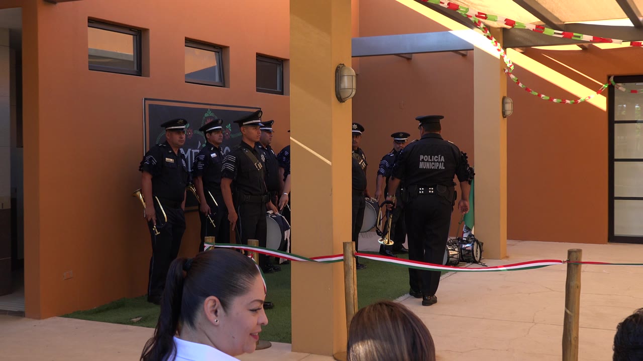Uniformed municipal police officers stand in a shaded exterior walkway in San Jose Del Cabo, with audience heads in the foreground and orange building walls behind them.