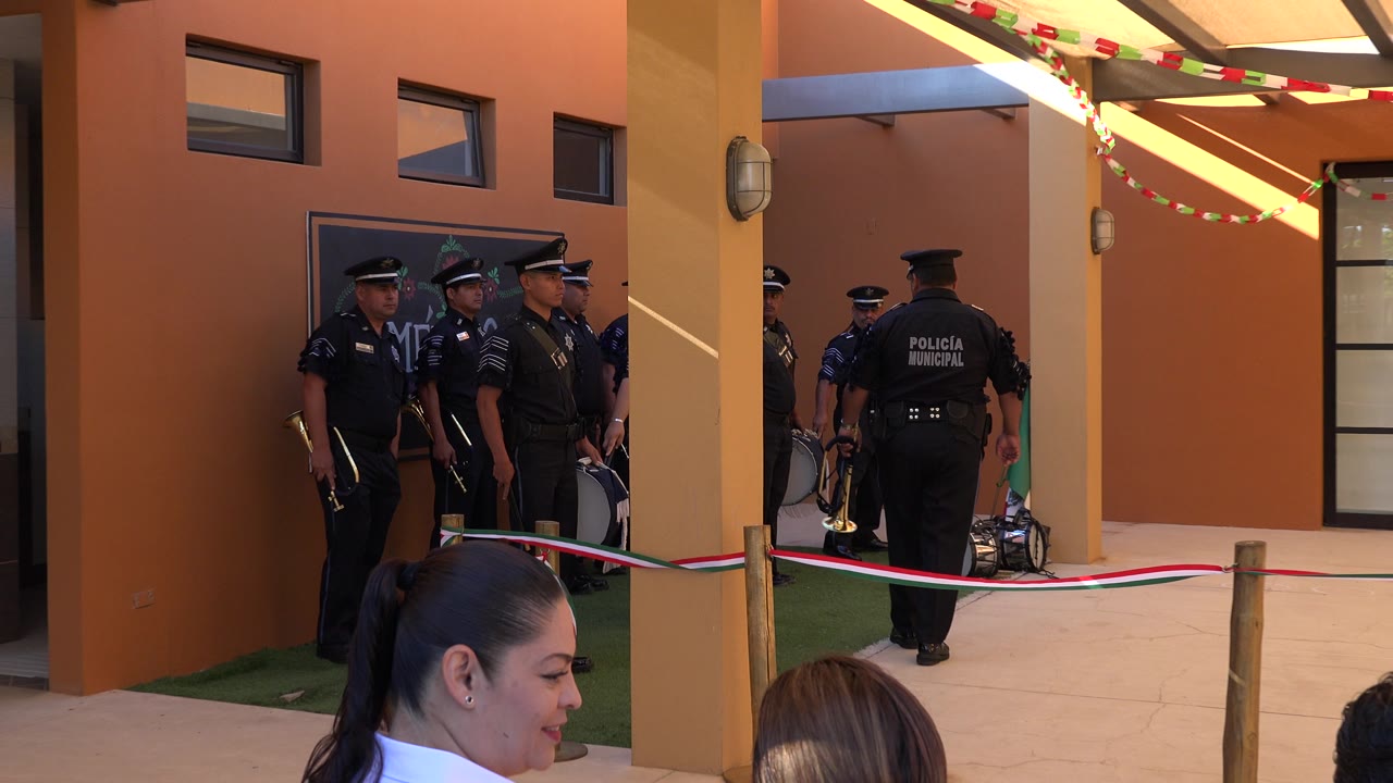 Several municipal police officers stand near a wall sign in San Jose Del Cabo, with columns, bunting, and a barrier separating them from foreground spectators.