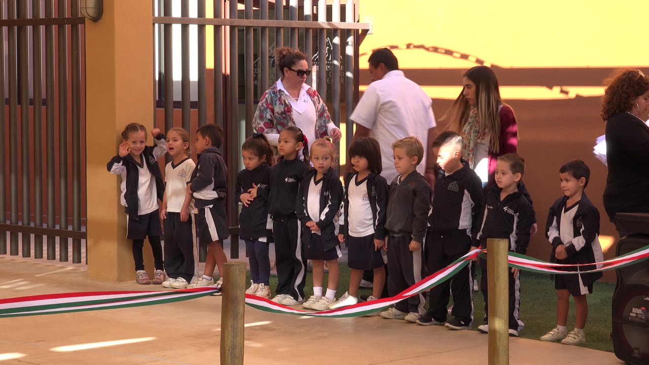 A row of children waits behind the ribbon in San Jose del Cabo, Mexico, with several adults supervising near the metal gate and warm-colored wall.