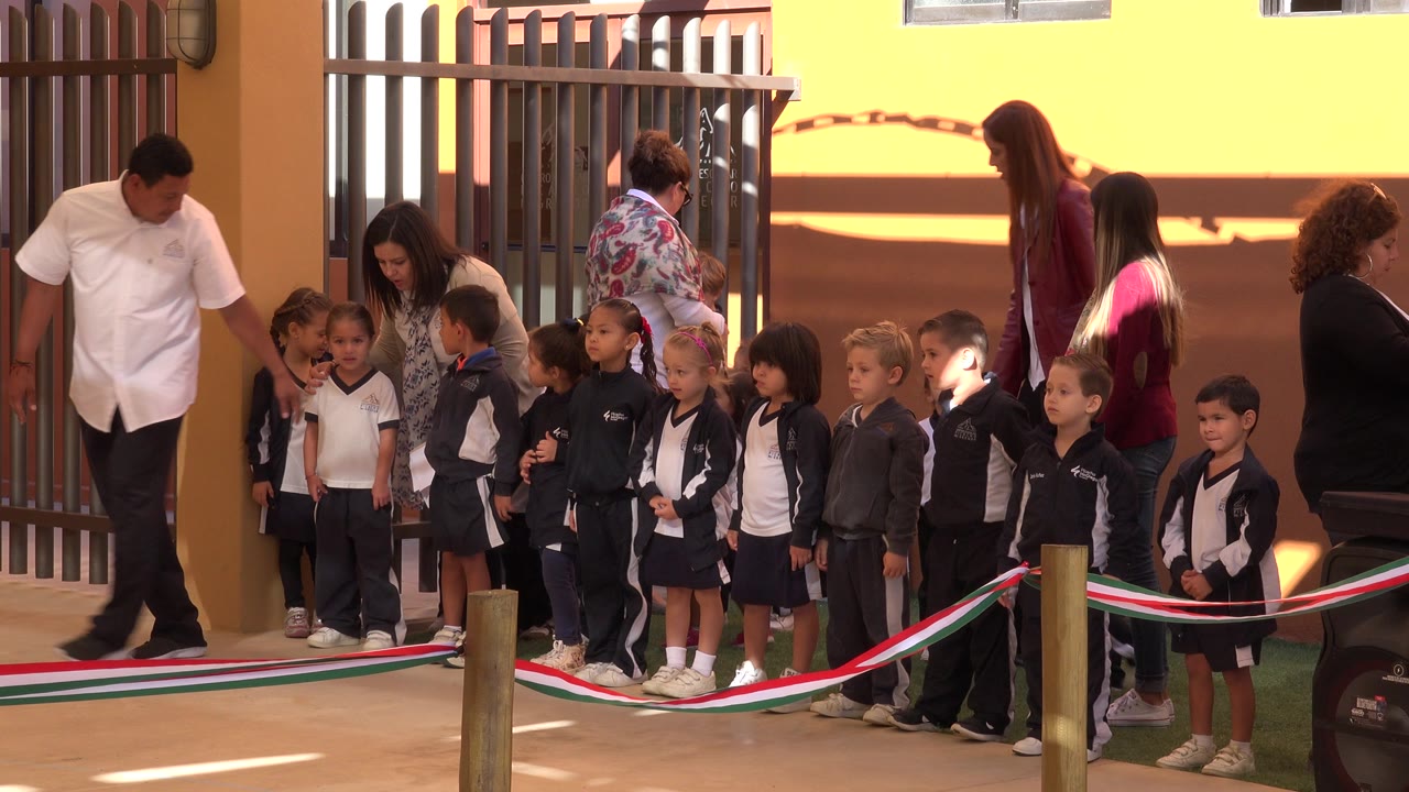In San Jose Del Cabo, Mexico, small children stand behind a red, white, and green rope while adults supervise beside a metal gate and warm painted wall.