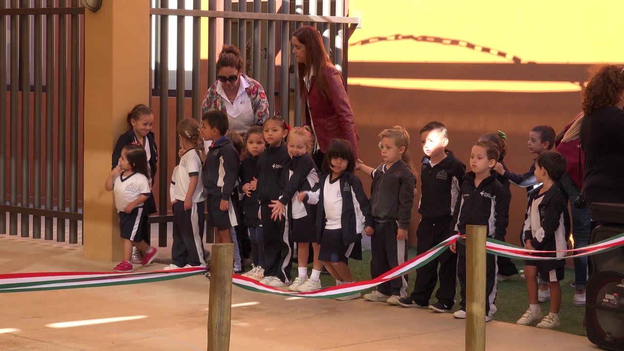 The line of children remains grouped near the gate in San Jose del Cabo, Mexico, with one adult leaning toward them and a rope barrier crossing the foreground.