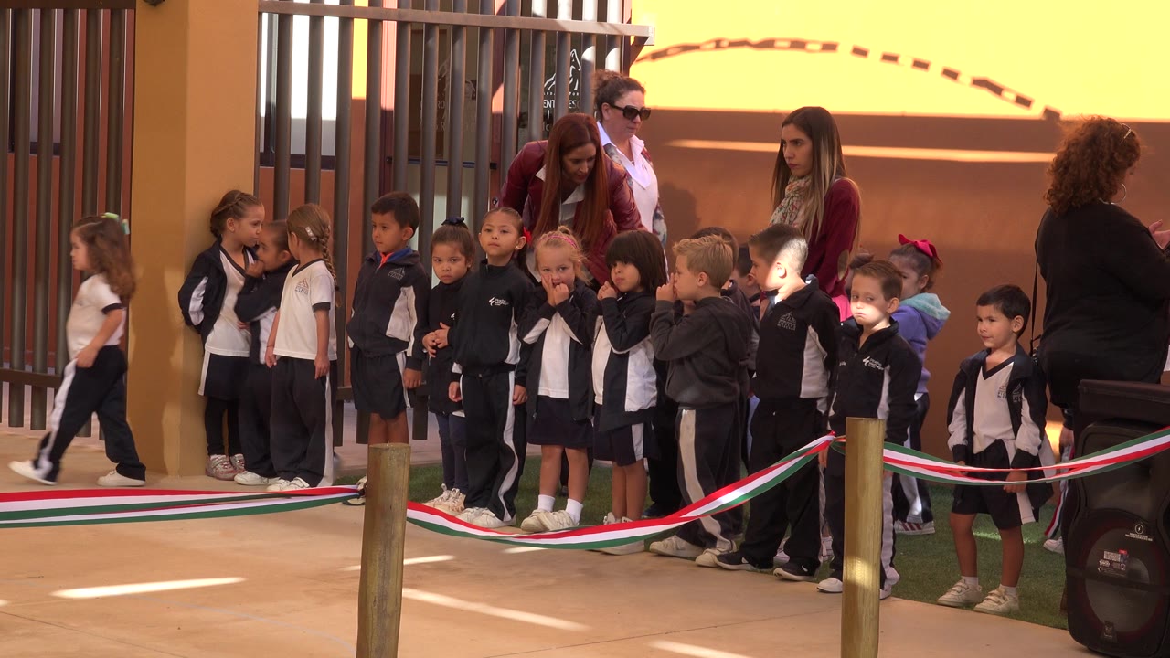 At 0:00, young children stand in a row outside in San Jose Del Cabo, Mexico, while adult women supervise near a yellow wall and metal gate.
