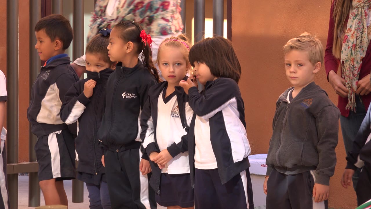 Uniformed kindergarten children stand in a loose line at a school in San Jose del Cabo, Mexico, with an adult partly visible behind them.