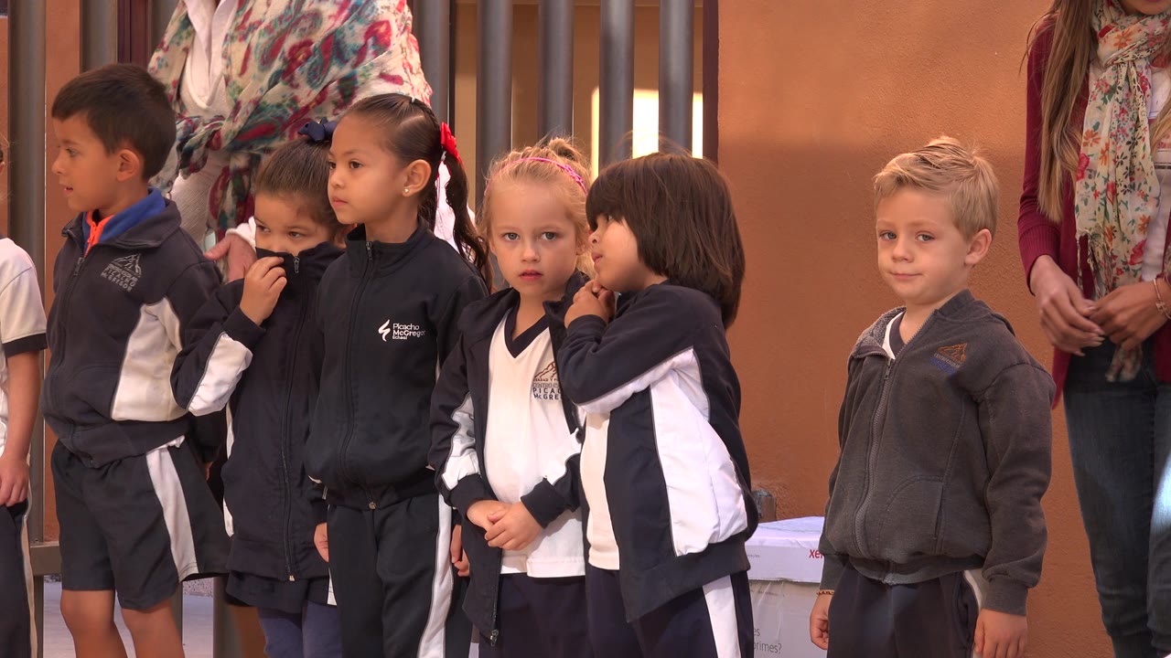 Several kindergarten-age children stand shoulder to shoulder in school uniforms, with warm walls and cropped adults in the background in San Jose del Cabo, Mexico.