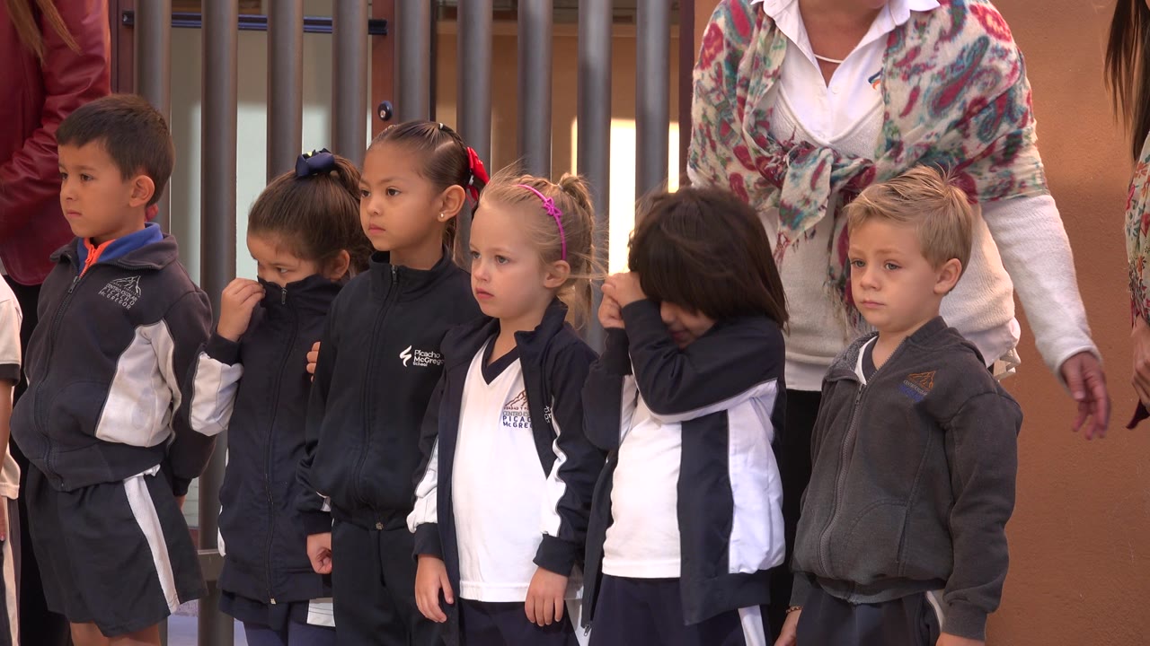 Children in school tracksuits stand beside a gated entrance with adults behind them.