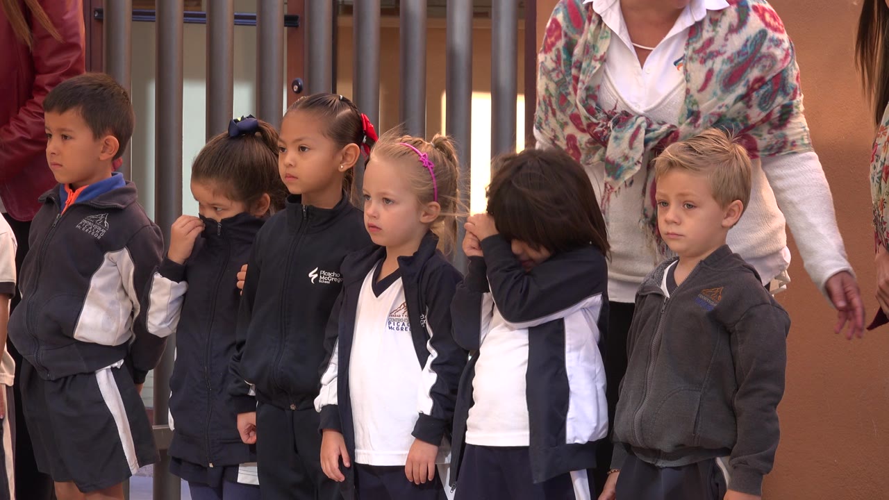 A row of young students in navy jackets and white shirts stands at a school entrance in San Jose del Cabo, Mexico, while an adult stands close behind them.