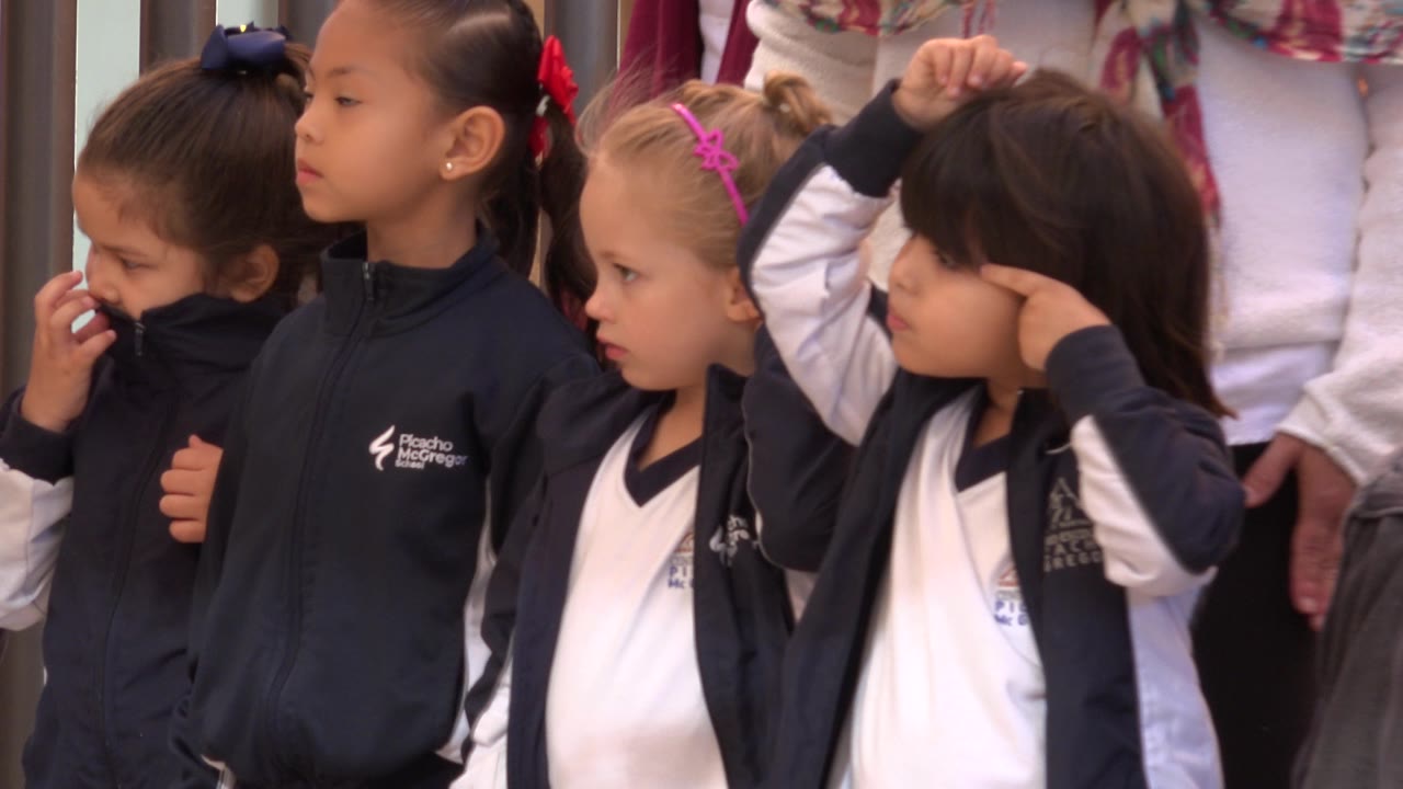 Four young girls in school uniforms stand together, one covering her nose.