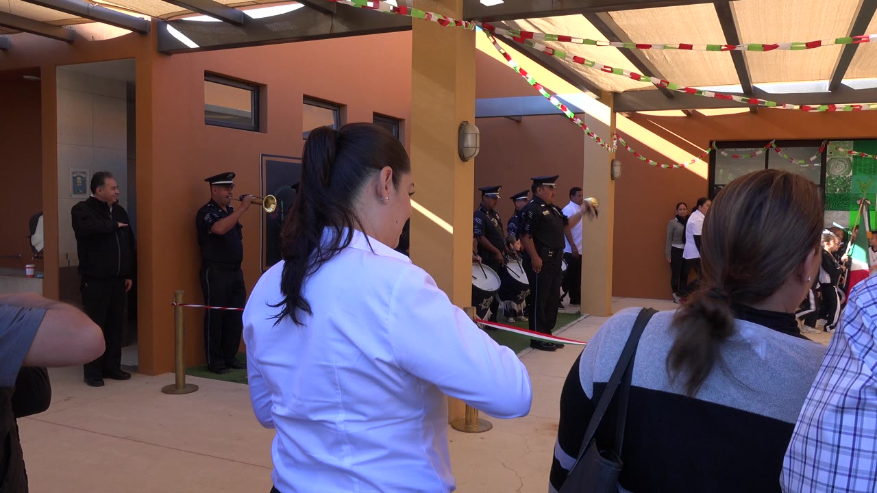 In San Jose del Cabo, Mexico, adults seen from behind watch uniformed musicians with brass and drums near an orange wall under a shaded roof.