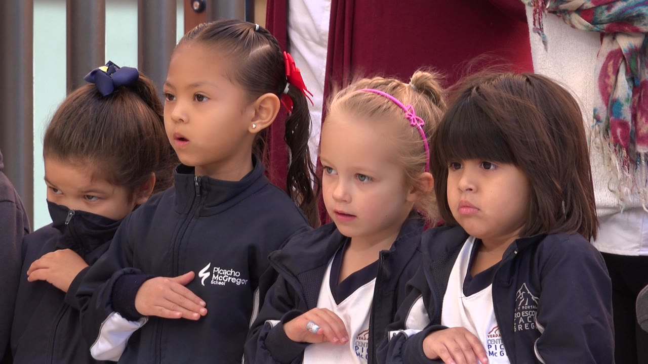 Four young girls in San Jose del Cabo stand shoulder to shoulder indoors, with crossed hands, hair bows, and red curtains behind them.
