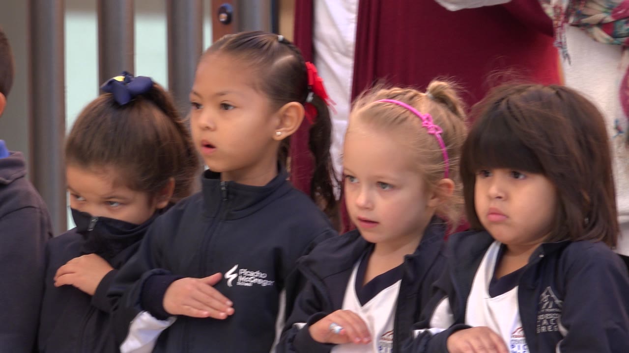 Girls in matching dark jackets stand indoors with adults behind them.